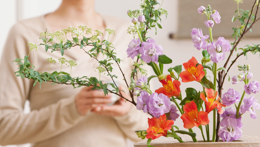 Close-up of vibrant orange and purple flowers arranged in a vase with green foliage, blurred person in beige sweater in background