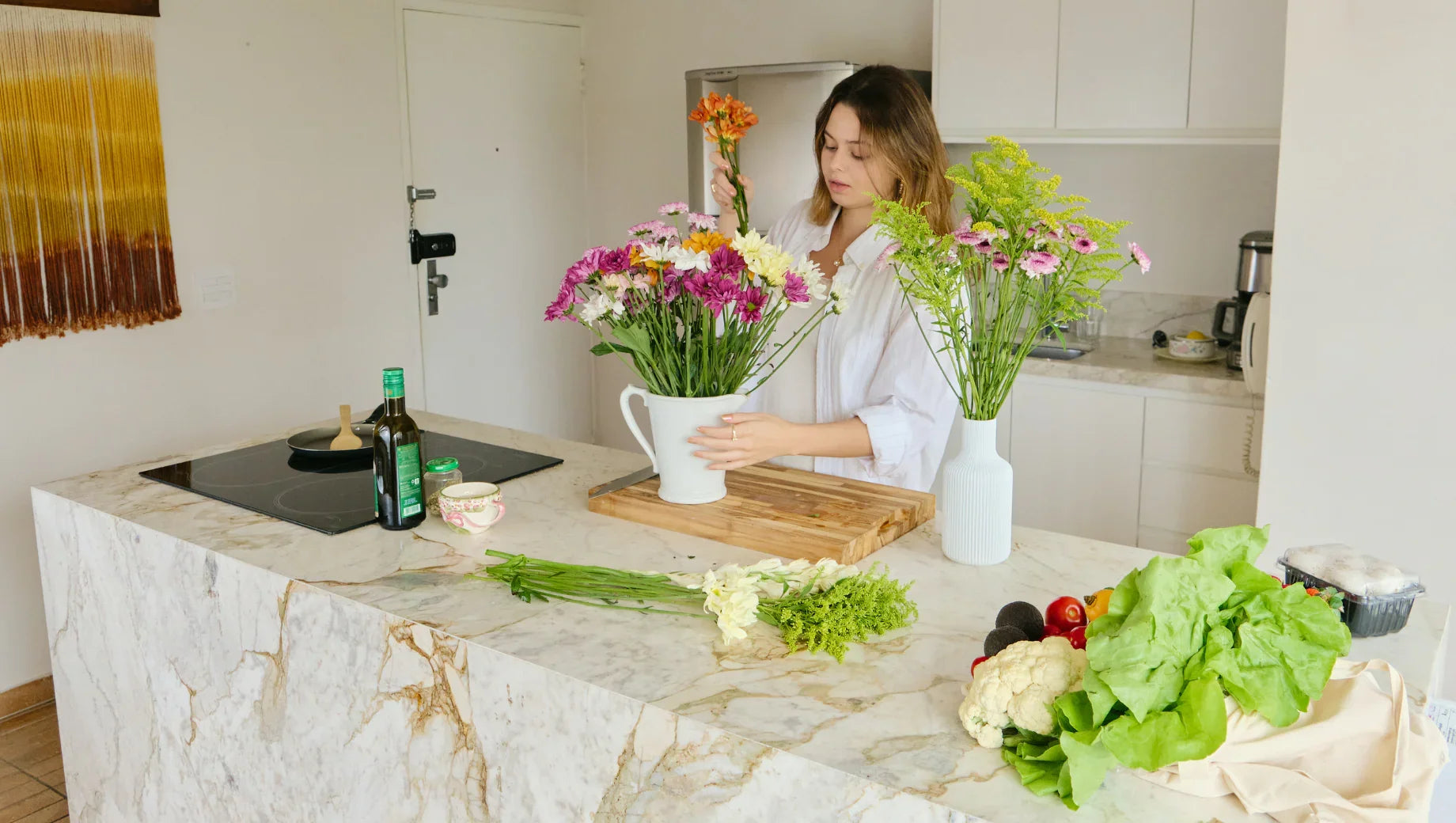 Woman arranging colorful flowers in white vase on marble kitchen island with fresh vegetables nearby
