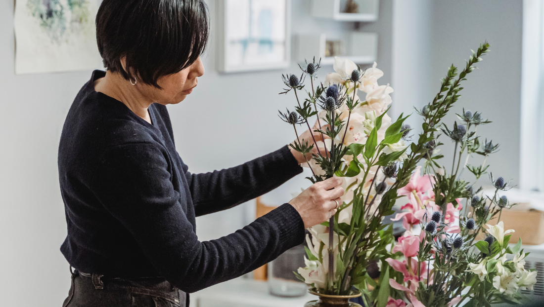 Woman arranging a mixed flower bouquet with green leaves and thistle flowers indoors