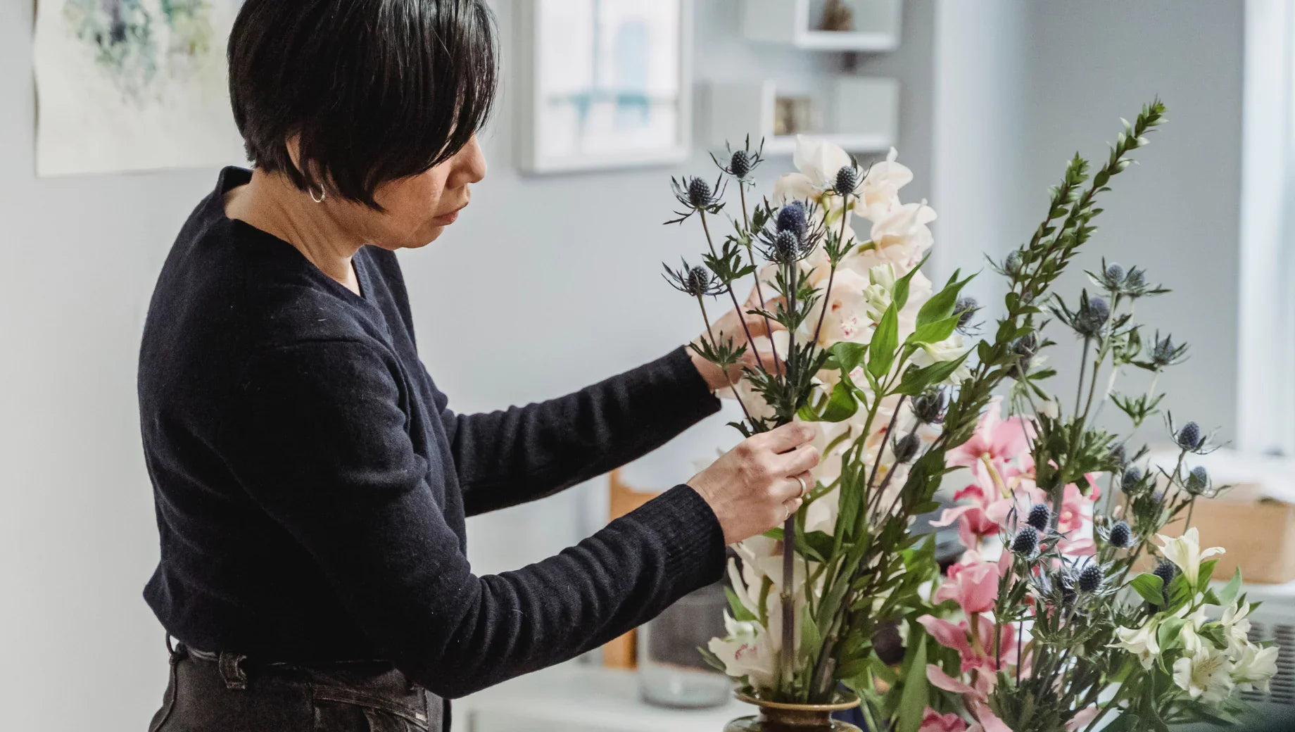 Woman arranging a mixed flower bouquet with green leaves and thistle flowers indoors