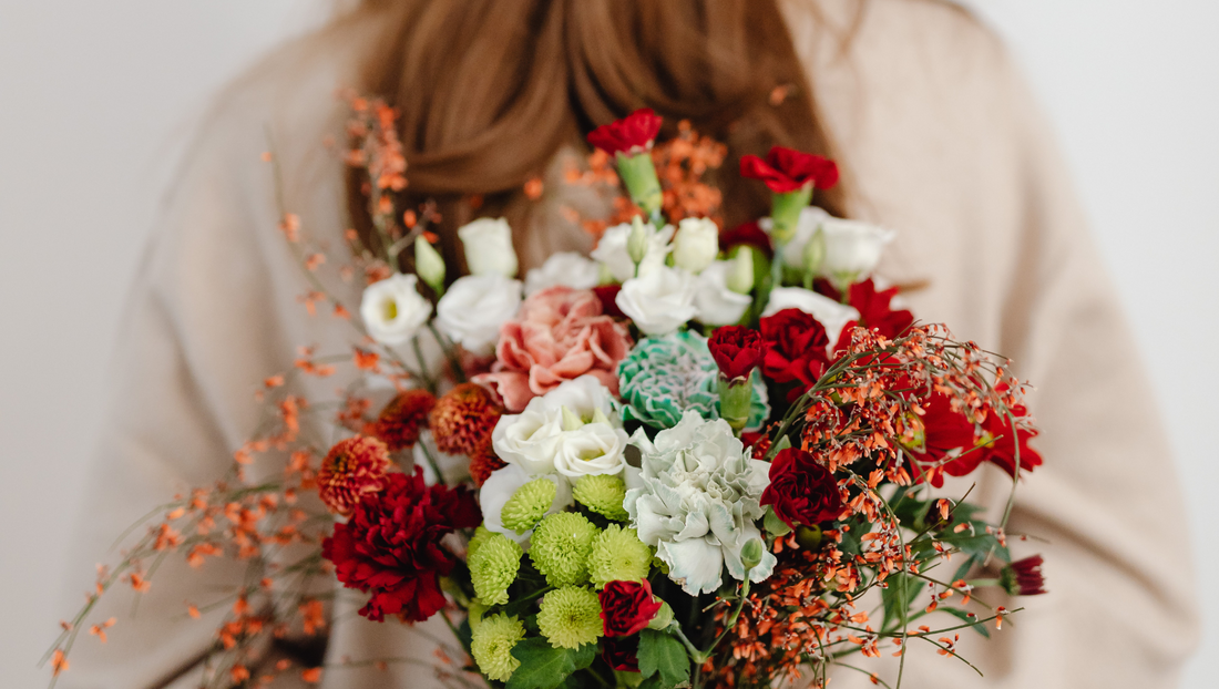 Person holding a vibrant bouquet of red, white, green, and peach flowers with delicate orange accents