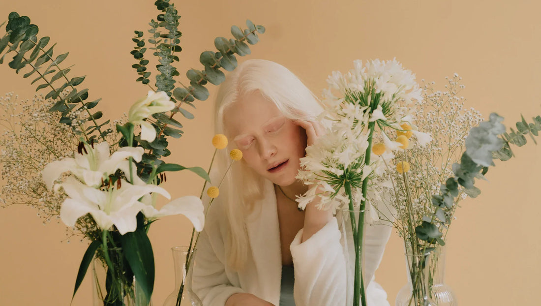 Albino woman in white robe surrounded by assorted white and yellow flowers in glass vases