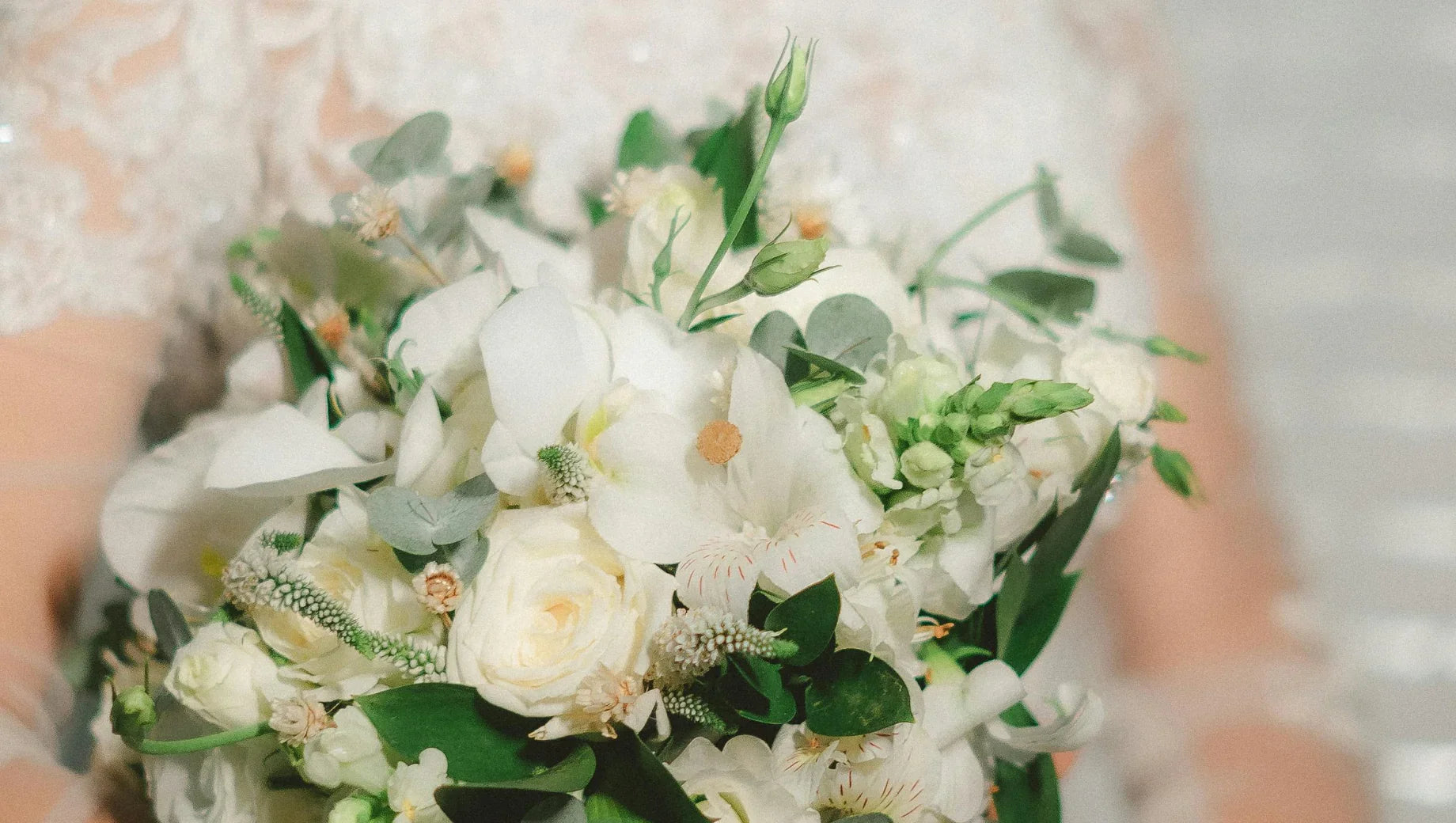 Close-up of a white bridal bouquet with roses, orchids, greenery, and lace wedding dress background