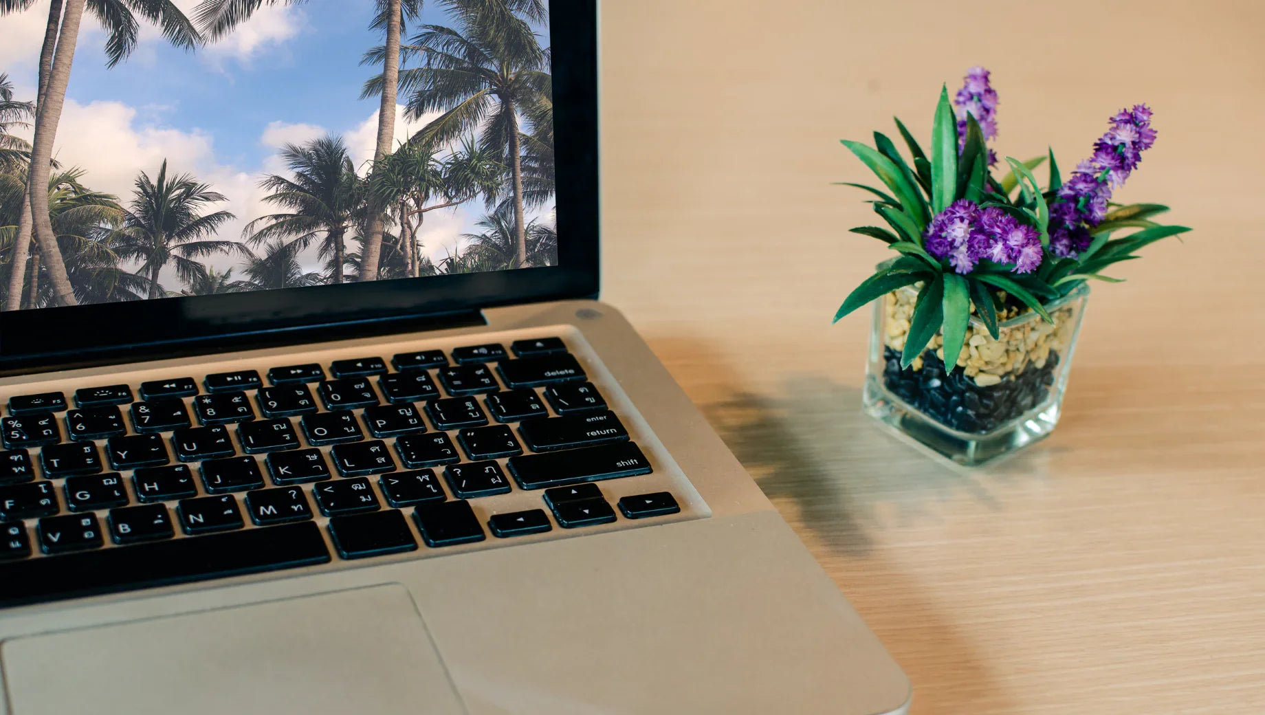 Laptop with palm tree wallpaper and small glass pot with purple flowers on wooden desk