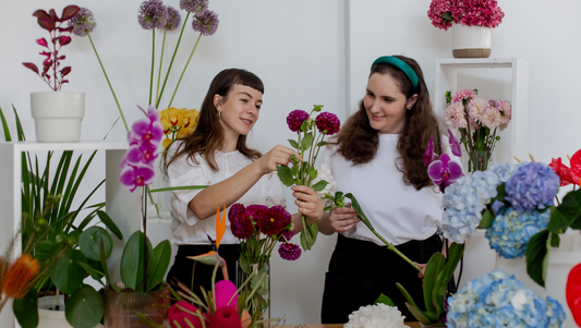 Two women arranging colorful flowers in a bright floral shop with orchids and hydrangeas