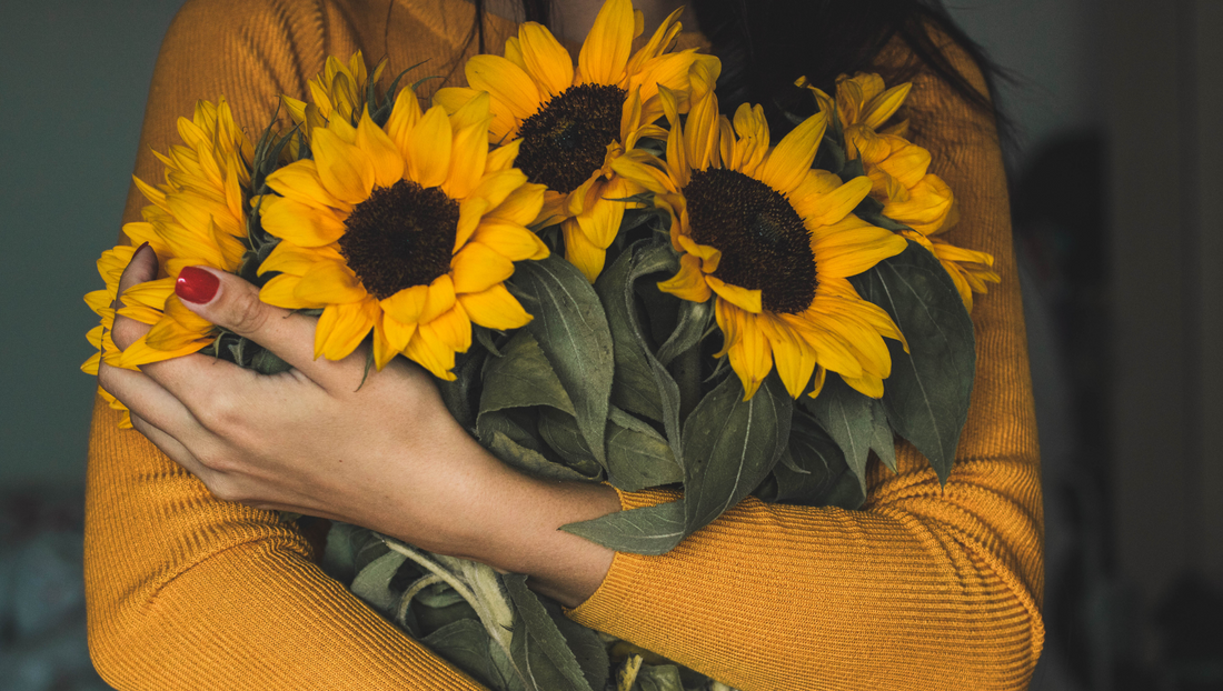 Person in mustard sweater holding a bouquet of bright yellow sunflowers with green leaves