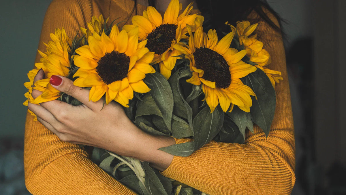 Person in mustard sweater holding a bouquet of bright yellow sunflowers with green leaves