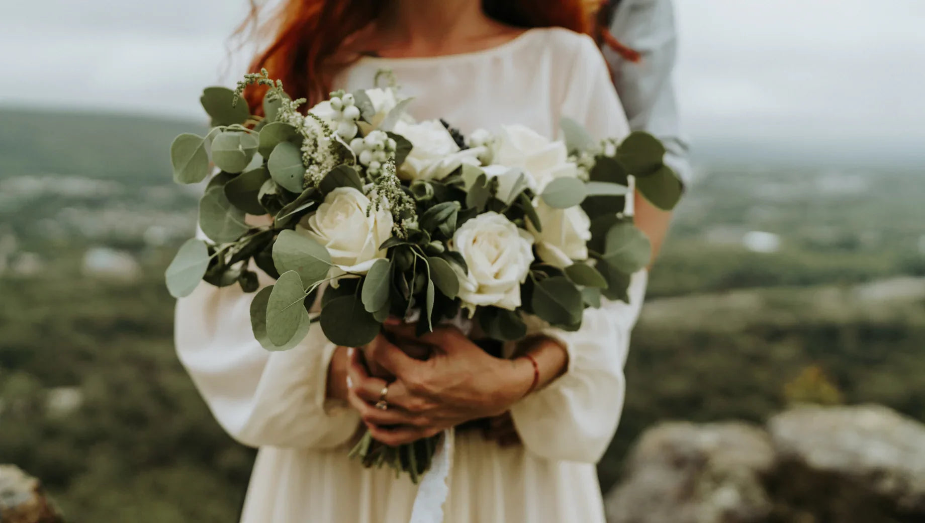 Couple holding white rose and eucalyptus wedding bouquet outdoors with landscape background