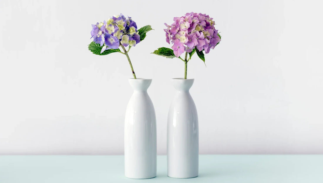 Two white ceramic vases each holding a single purple hydrangea flower with green leaves on light blue surface