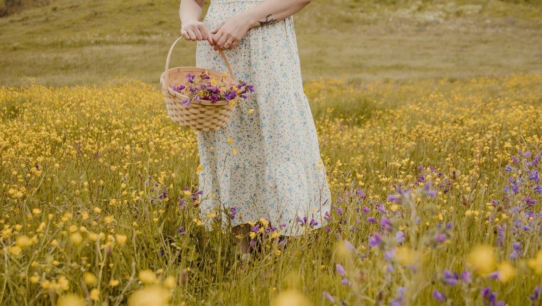 Woman in floral dress holding basket of purple and yellow wildflowers in a blooming meadow