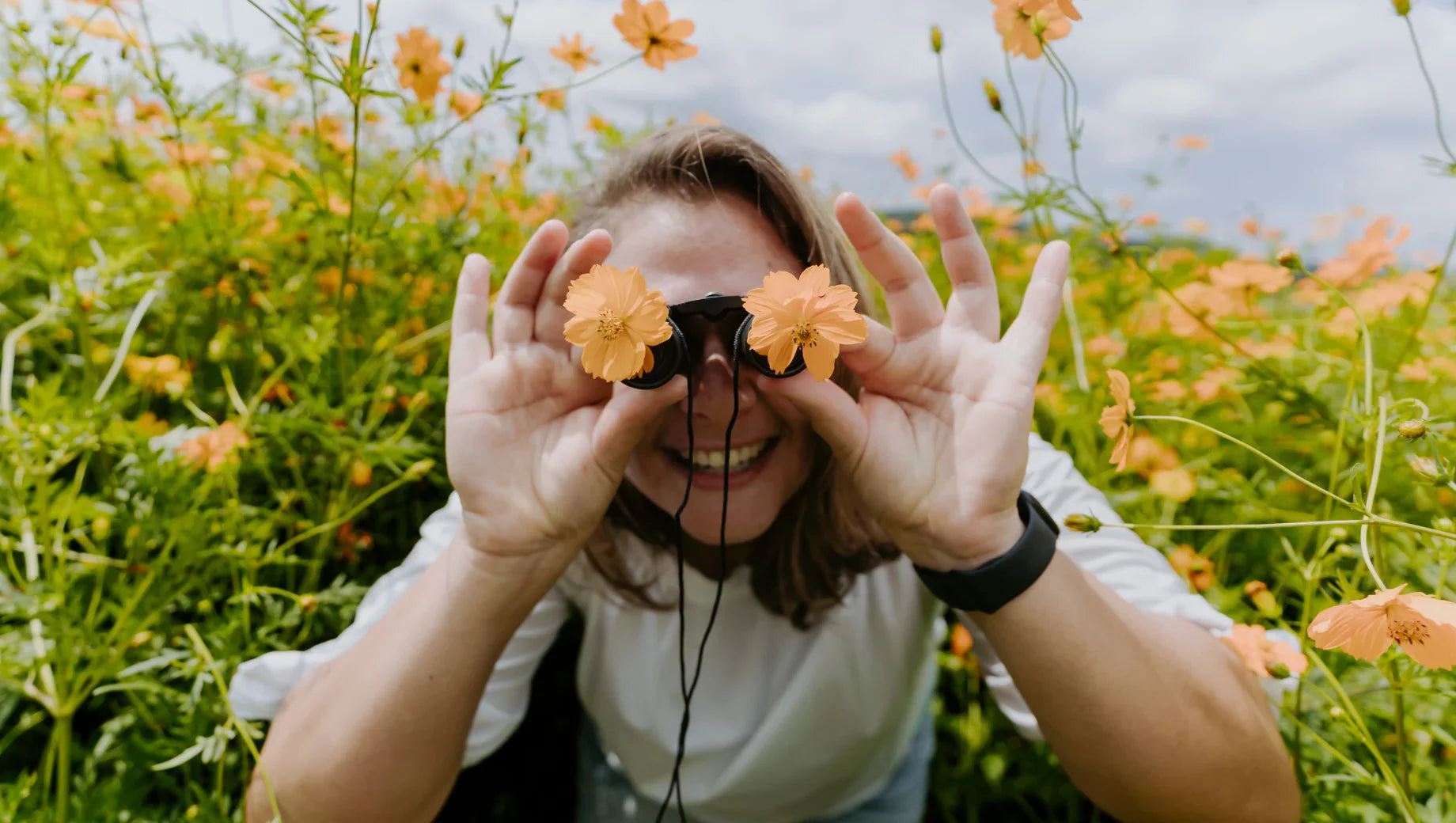 Woman in white shirt smiling in orange flower field holding binoculars with flowers in front