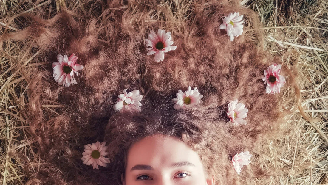 Woman lying on hay with curly hair adorned with pink daisies in rustic outdoor setting