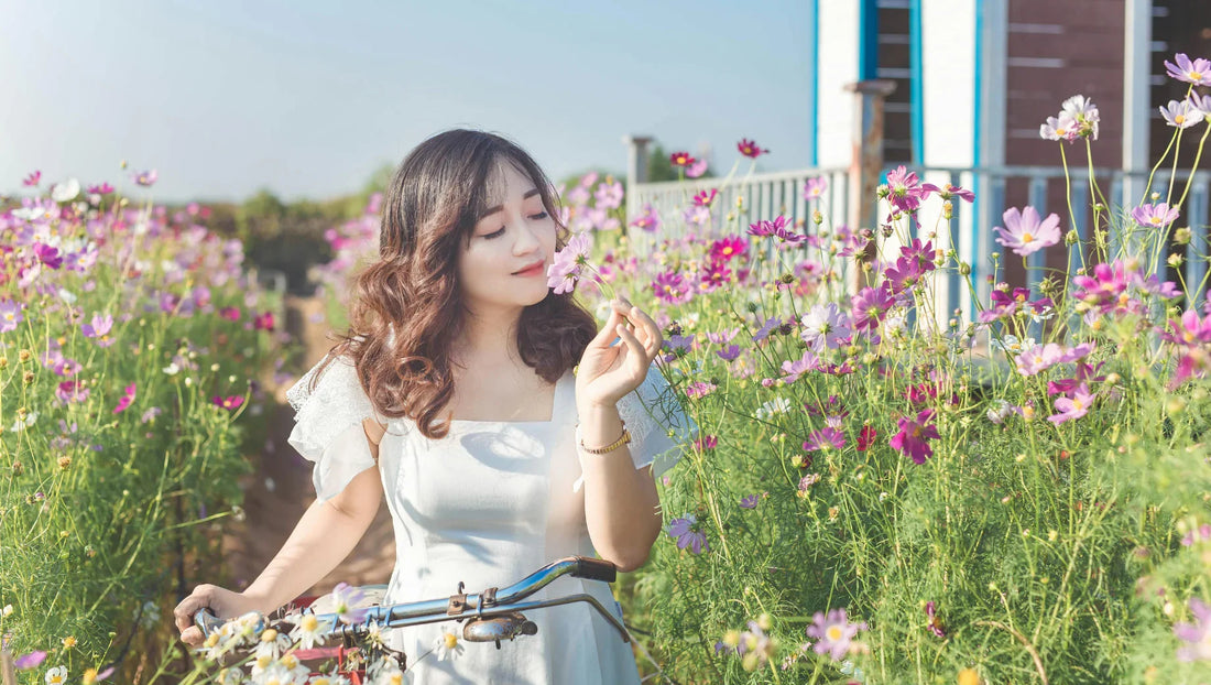 Young woman in white dress enjoying pink flowers in a sunny flower garden holding bicycle