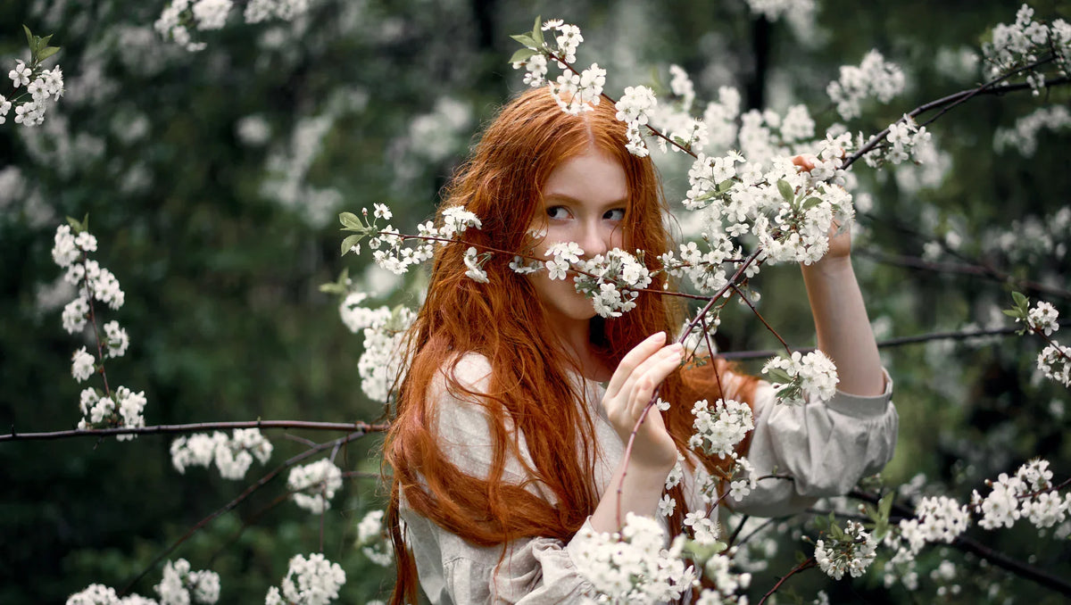 Red-haired woman in beige dress surrounded by blooming white flowers on tree branches