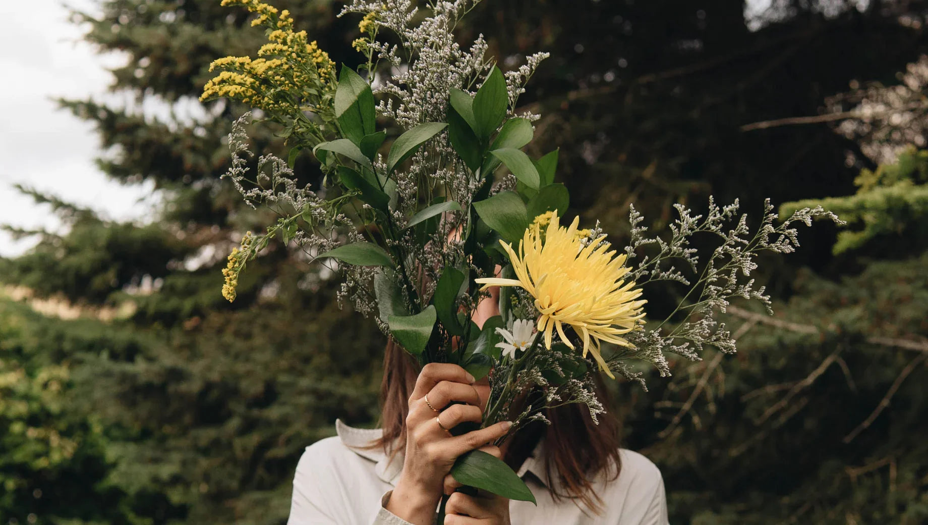 Person holding a bouquet of mixed flowers with yellow chrysanthemum outdoors among green trees
