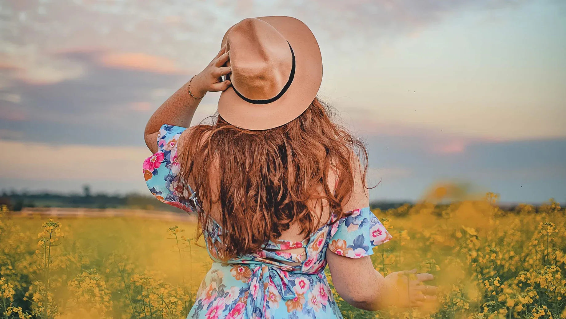 Woman with red hair wearing a floral dress and tan hat in a yellow flower field at sunset
