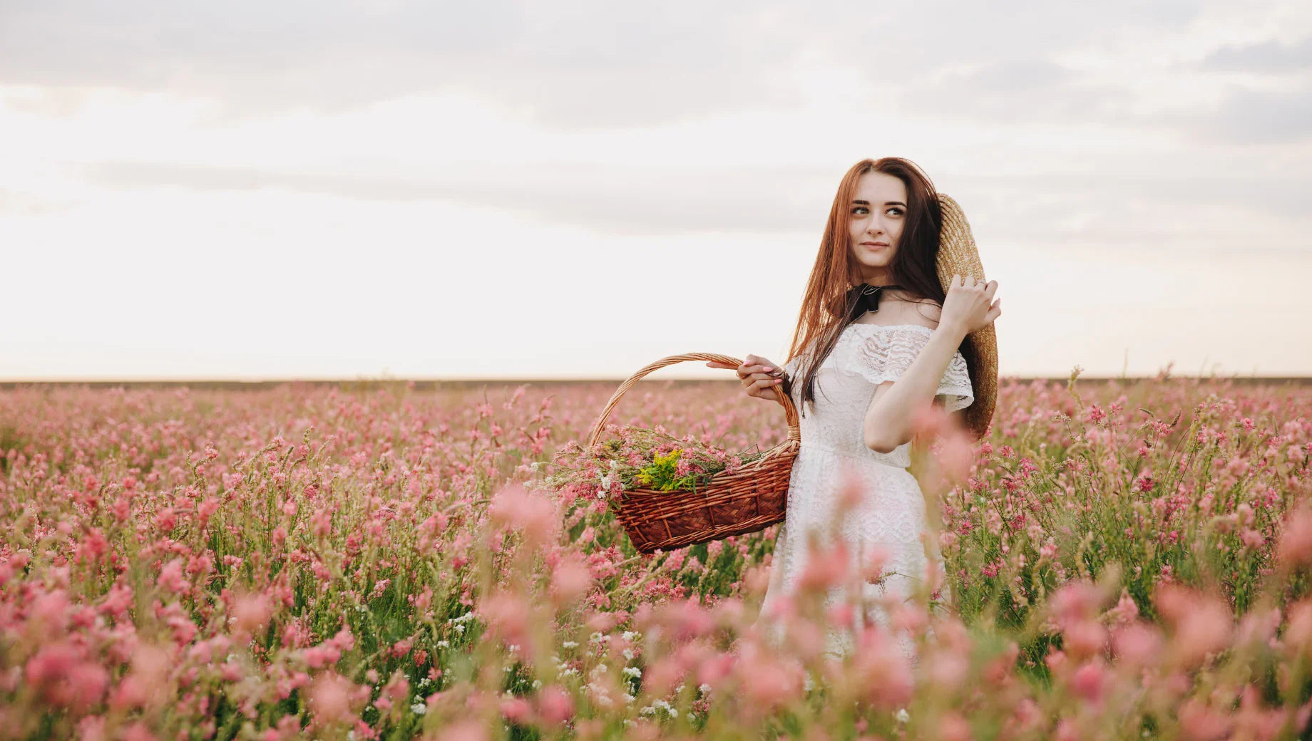 Young woman in white dress holding basket with flowers and straw hat in a pink flower field at sunset